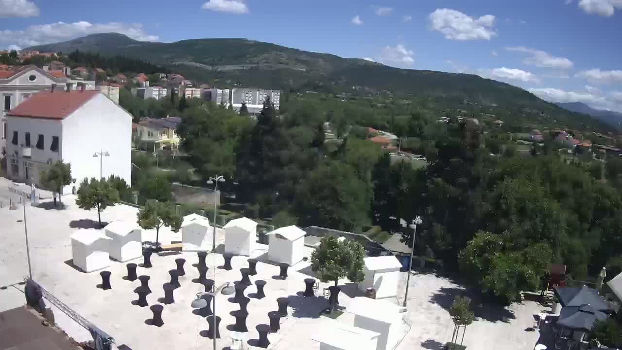 A high-angle view shows a town square with small white structures and black-clothed tables under a partly cloudy sky, overlooking a hilly landscape with lush green vegetation.