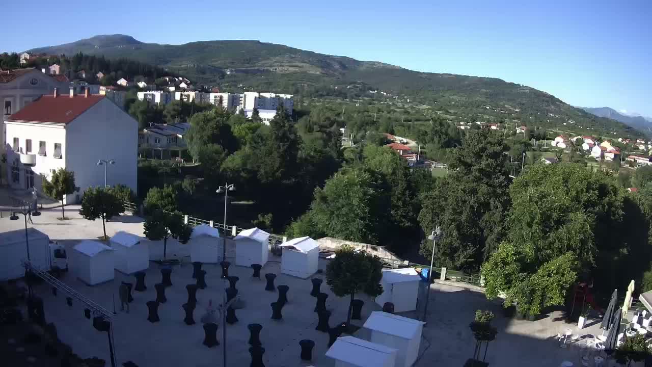 A town square with numerous small white structures and black tables, situated next to a river and surrounded by lush green trees and mountains under a clear blue sky.