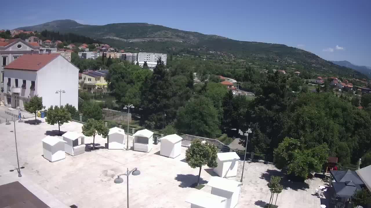 A town square with several small white kiosks is situated below a hill covered in lush green trees and houses under a clear blue sky.