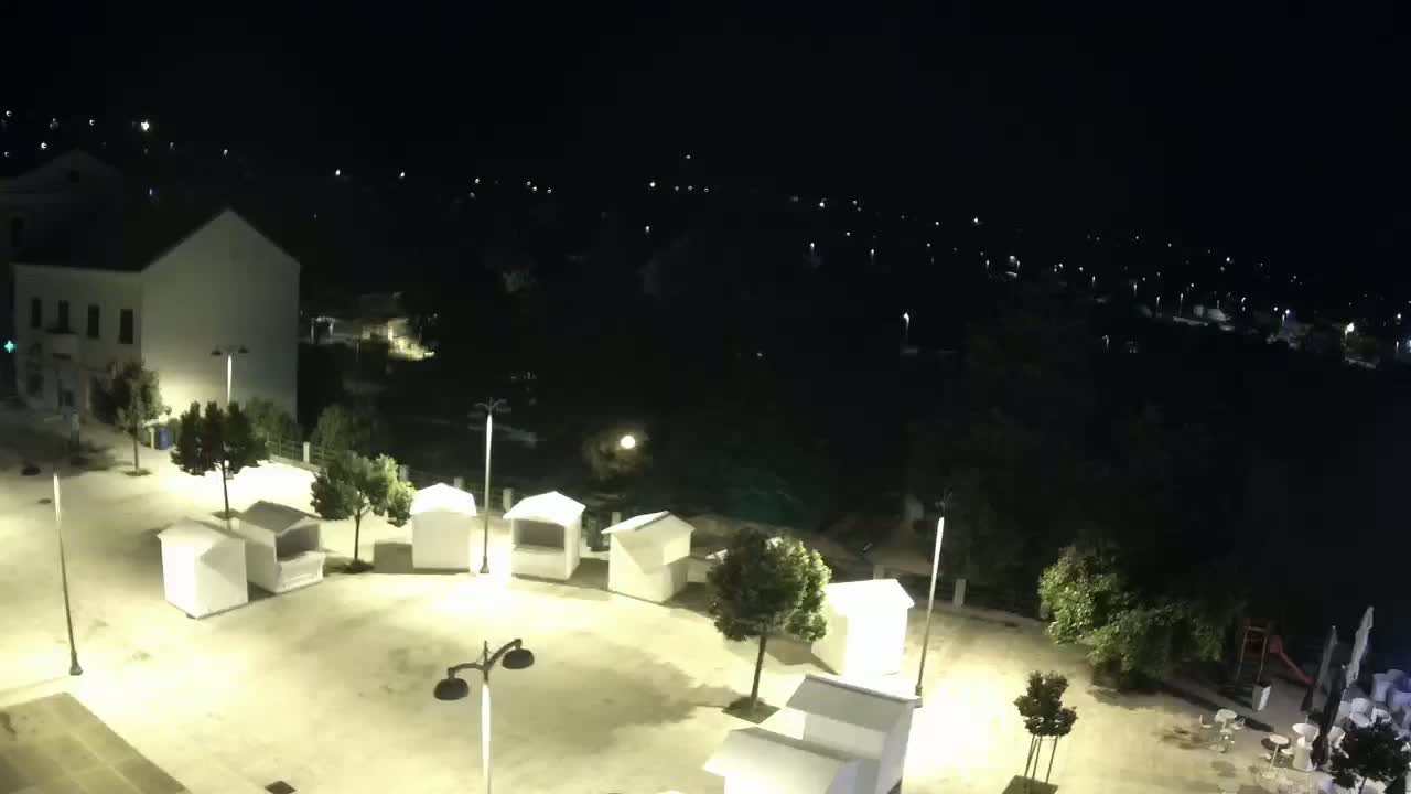An empty, illuminated plaza at night with several small white kiosks, surrounded by trees and buildings, overlooks a dark body of water.