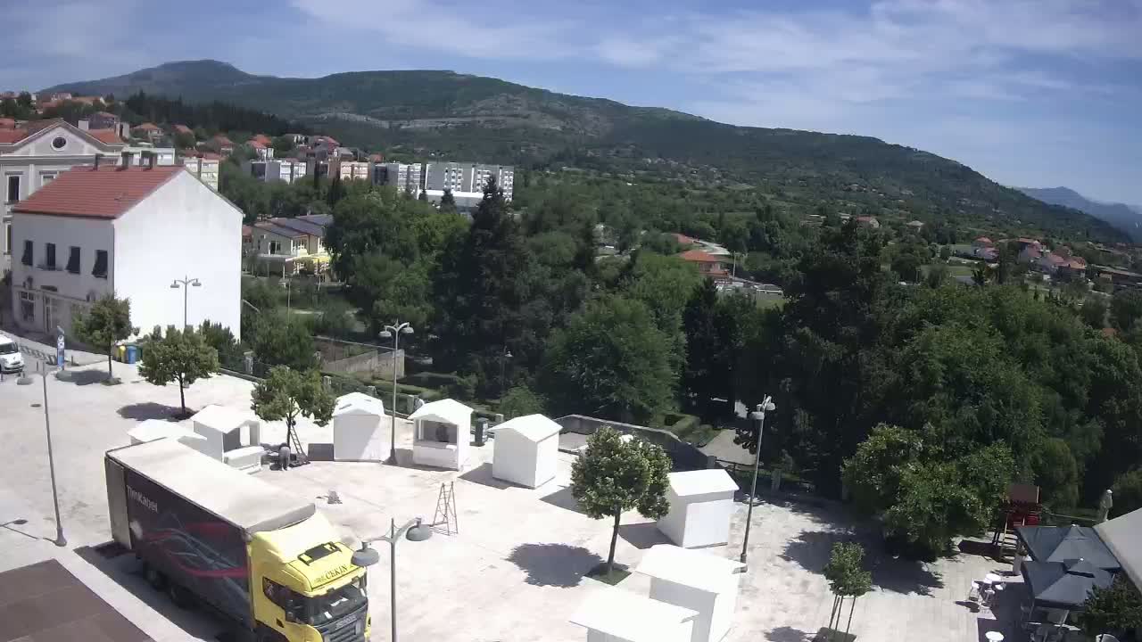 A town square with several small white structures, a yellow truck, and surrounding buildings, under a sunny sky with a mountain in the background.