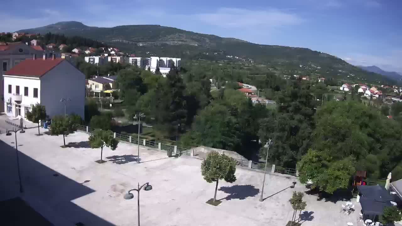 A sunny day view of a town square with several trees and buildings, overlooking a lush green valley and mountains in the distance.