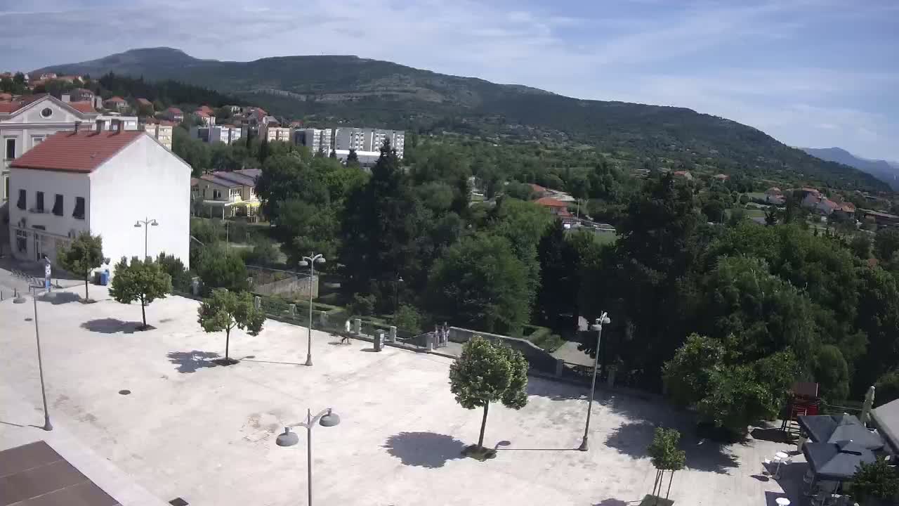 An empty town square with trees and lampposts is situated in front of buildings and a lush, green hillside under a mostly sunny sky.