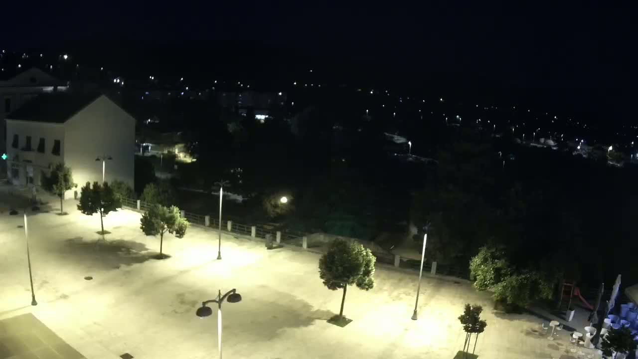 A nighttime, high-angle view of an empty town square illuminated by streetlights, with buildings and trees visible in the background under a clear, dark sky.