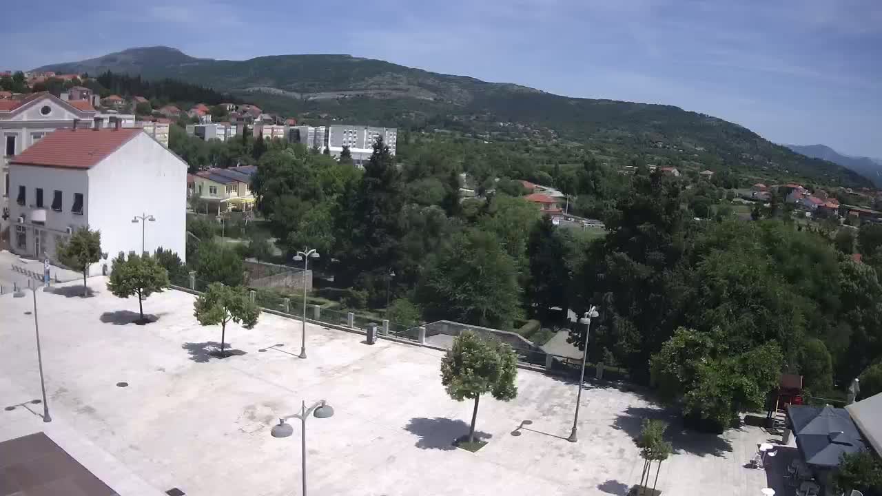 A mostly empty paved square with a few trees and lampposts is situated in front of buildings, overlooking a lush green hillside under a clear blue sky.