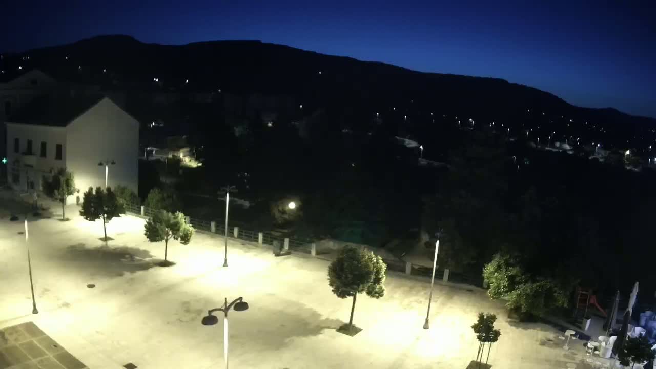A nighttime, high-angle view shows an empty plaza with several trees and lampposts, illuminated against a backdrop of dark hills and a dimly lit town.