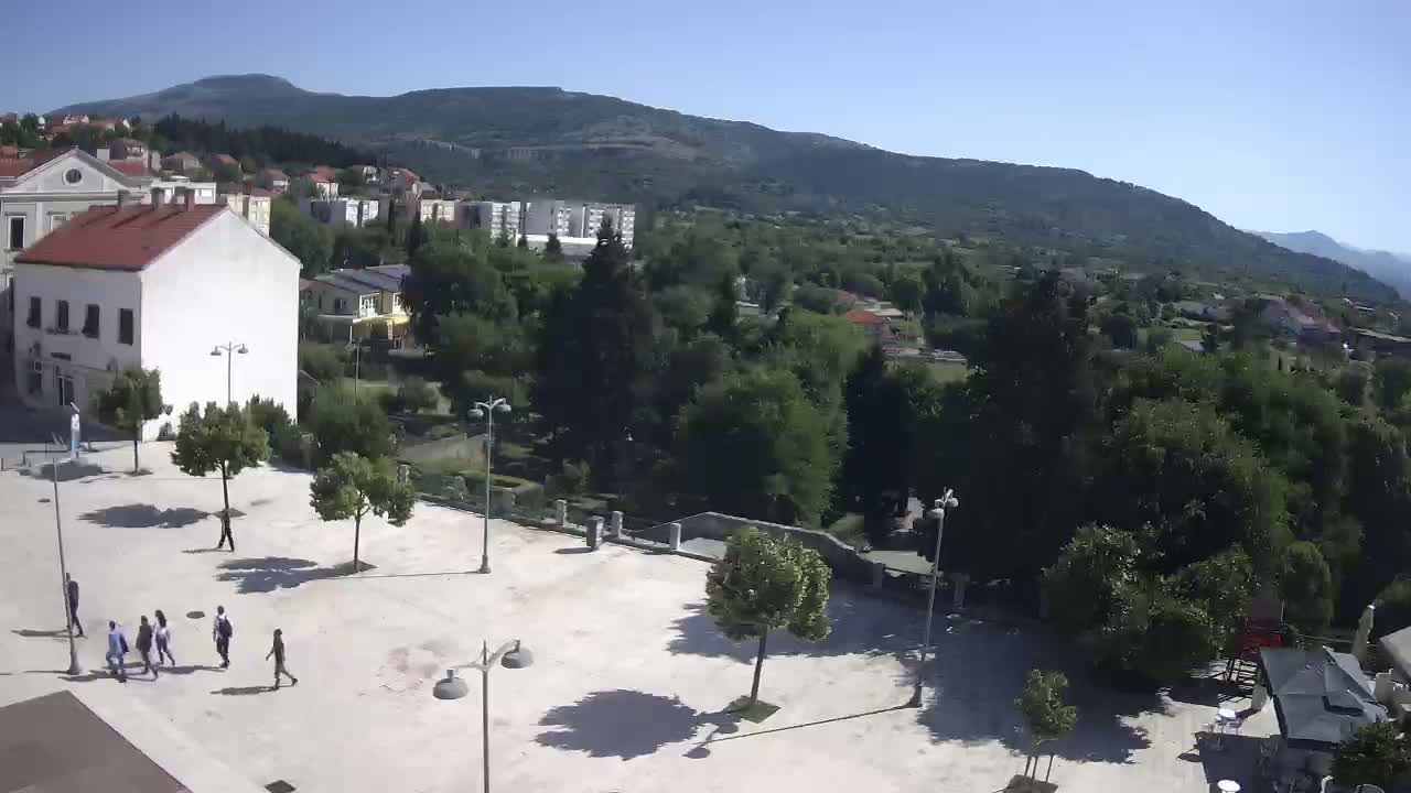 A mostly sunny outdoor scene shows a town square with several people walking, a few buildings, and a large expanse of green trees and hills in the background.