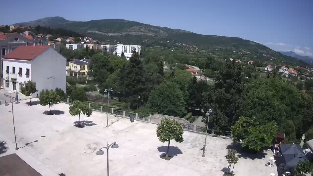 A paved town square with a few small trees and lampposts, situated near buildings and lush green hills under a clear, sunny sky.