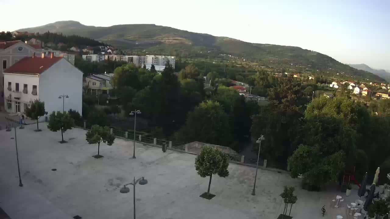 An elevated view of a town square with several trees and buildings, under a clear sky, overlooking a hilly landscape.