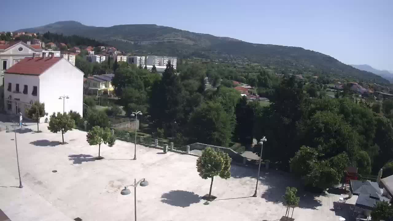 A mostly empty town square with several trees and lampposts, overlooks a verdant hillside under a clear, sunny sky.