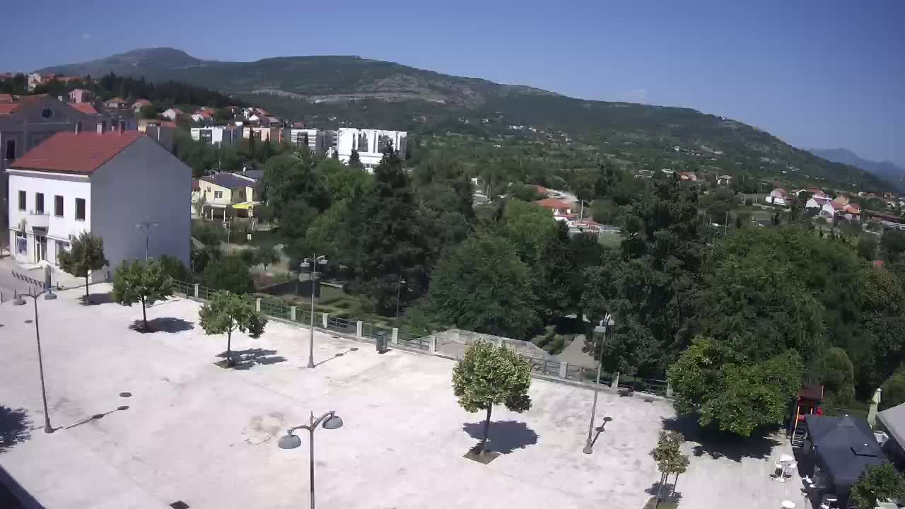 An empty, light-colored plaza with a few trees and lampposts is situated in front of a white building, overlooking a verdant hillside town under a clear, sunny sky.