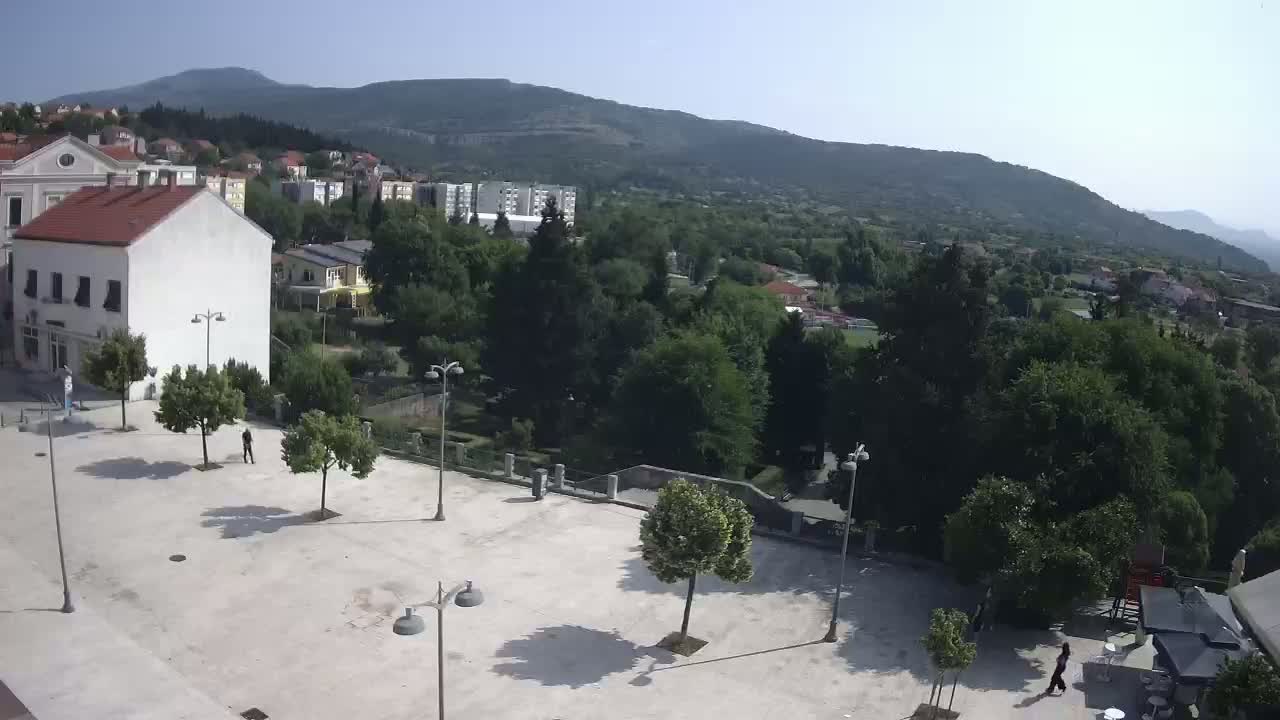 An empty town square with a few people and trees, overlooks a lush green hillside under a clear, sunny sky.