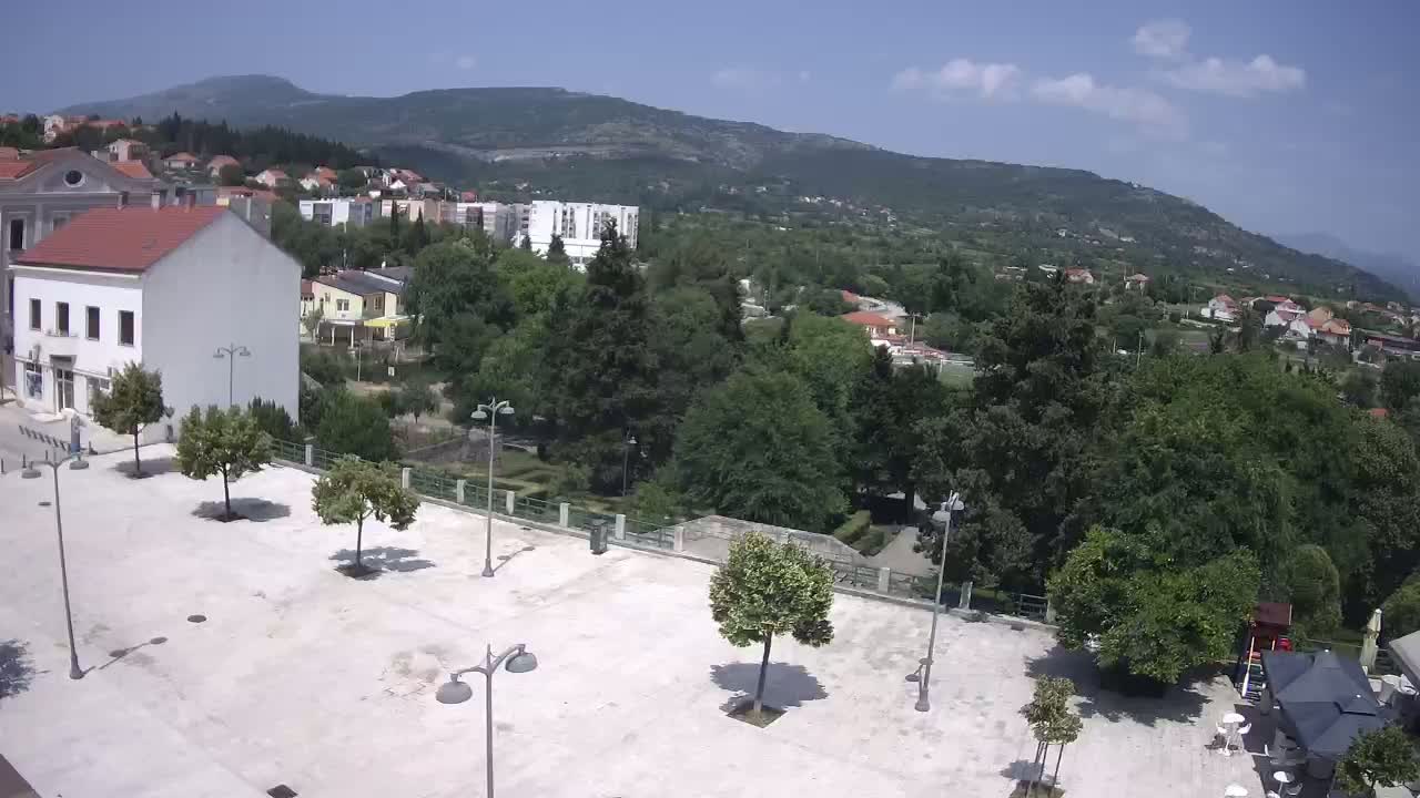 An empty town square with a few trees and lampposts is situated in front of buildings and a hillside covered in lush green vegetation under a mostly sunny sky.