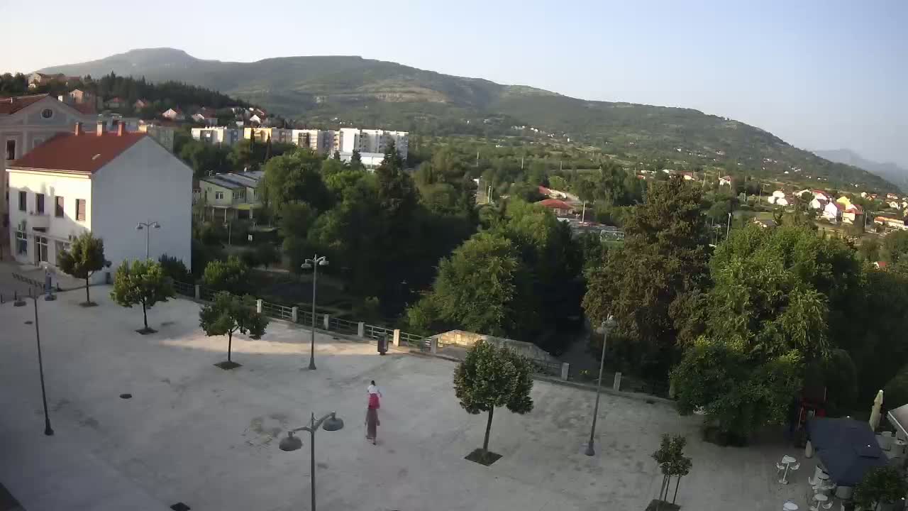 A paved town square with a few people and trees, overlooking a green valley and distant mountains under a clear sky.