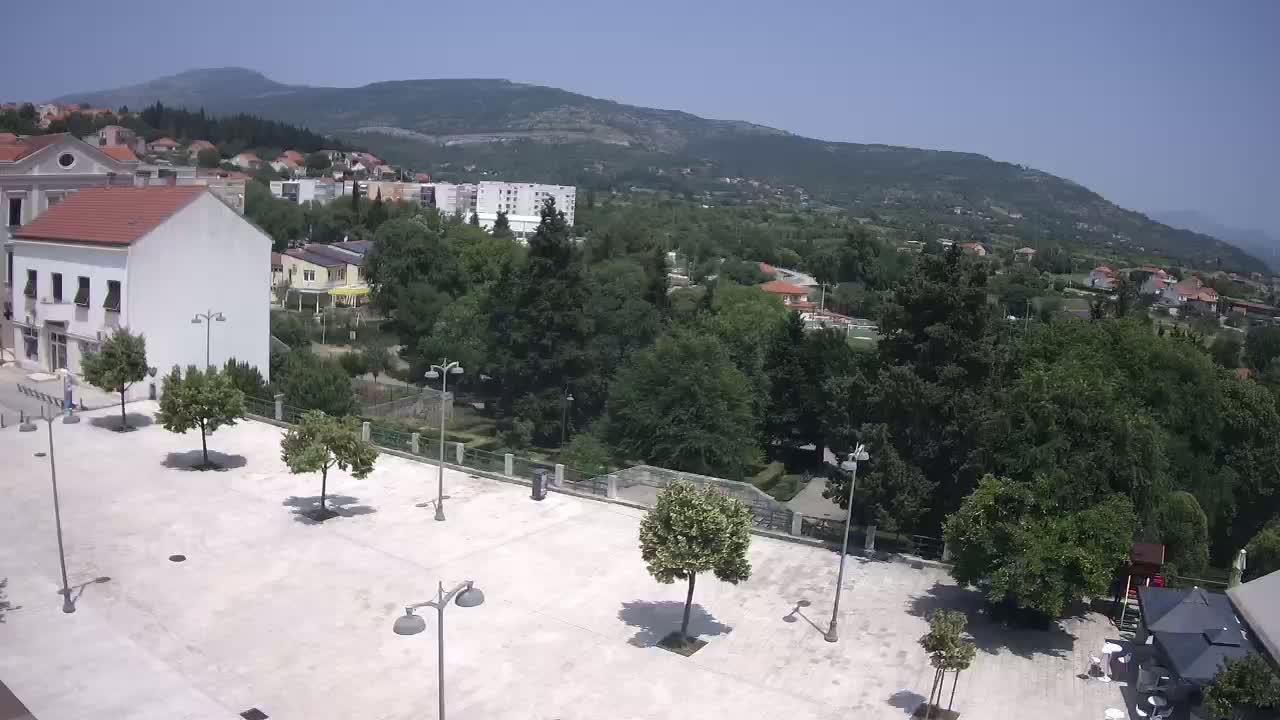 A high-angle view of a town square on a sunny day, with several trees and buildings surrounding a paved area, and mountains in the background.