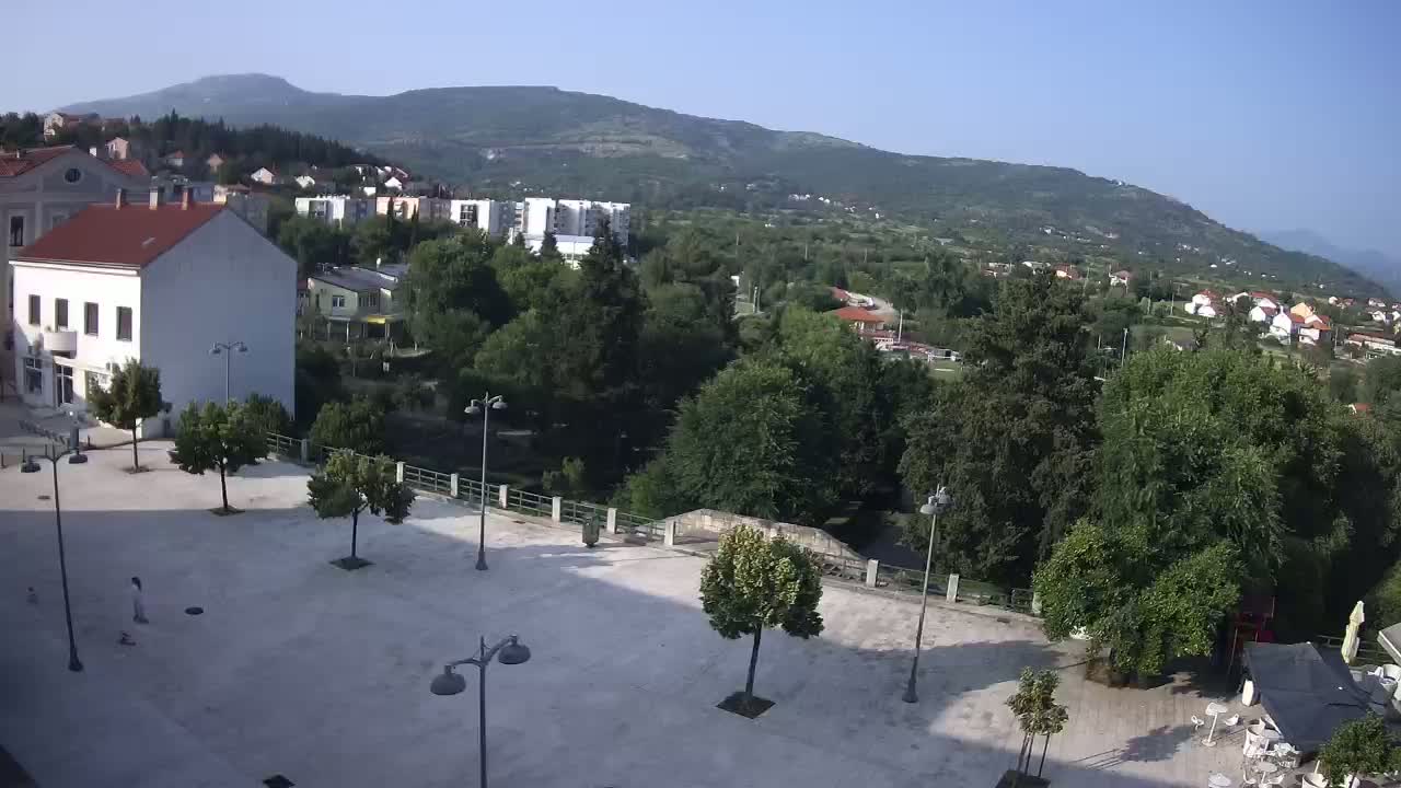 A mostly empty town square with several trees and lampposts, overlooks a lush green valley and distant mountains under a clear, sunny sky.