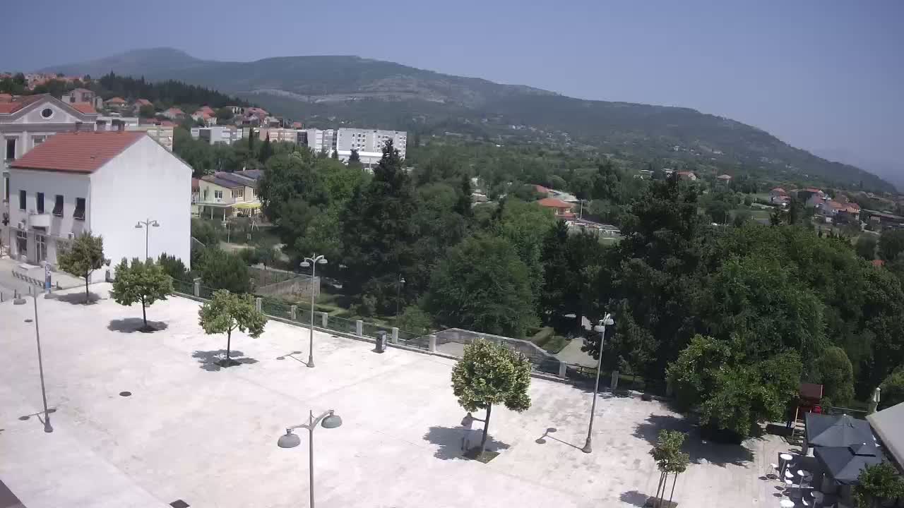 A paved town square with several trees and lampposts is situated in front of buildings, overlooking a hillside covered with lush green vegetation and houses under a clear, sunny sky.