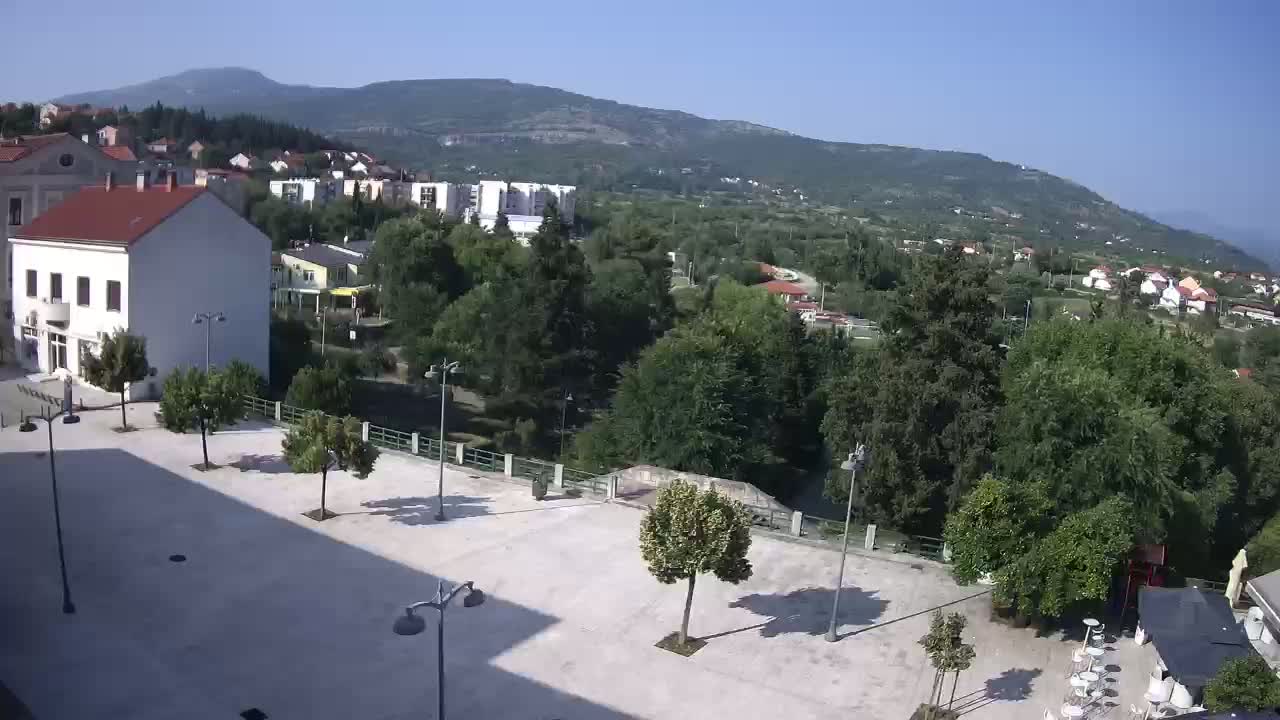 An empty town square with a few trees and lampposts, overlooked by buildings and a verdant hillside under a clear, sunny sky.