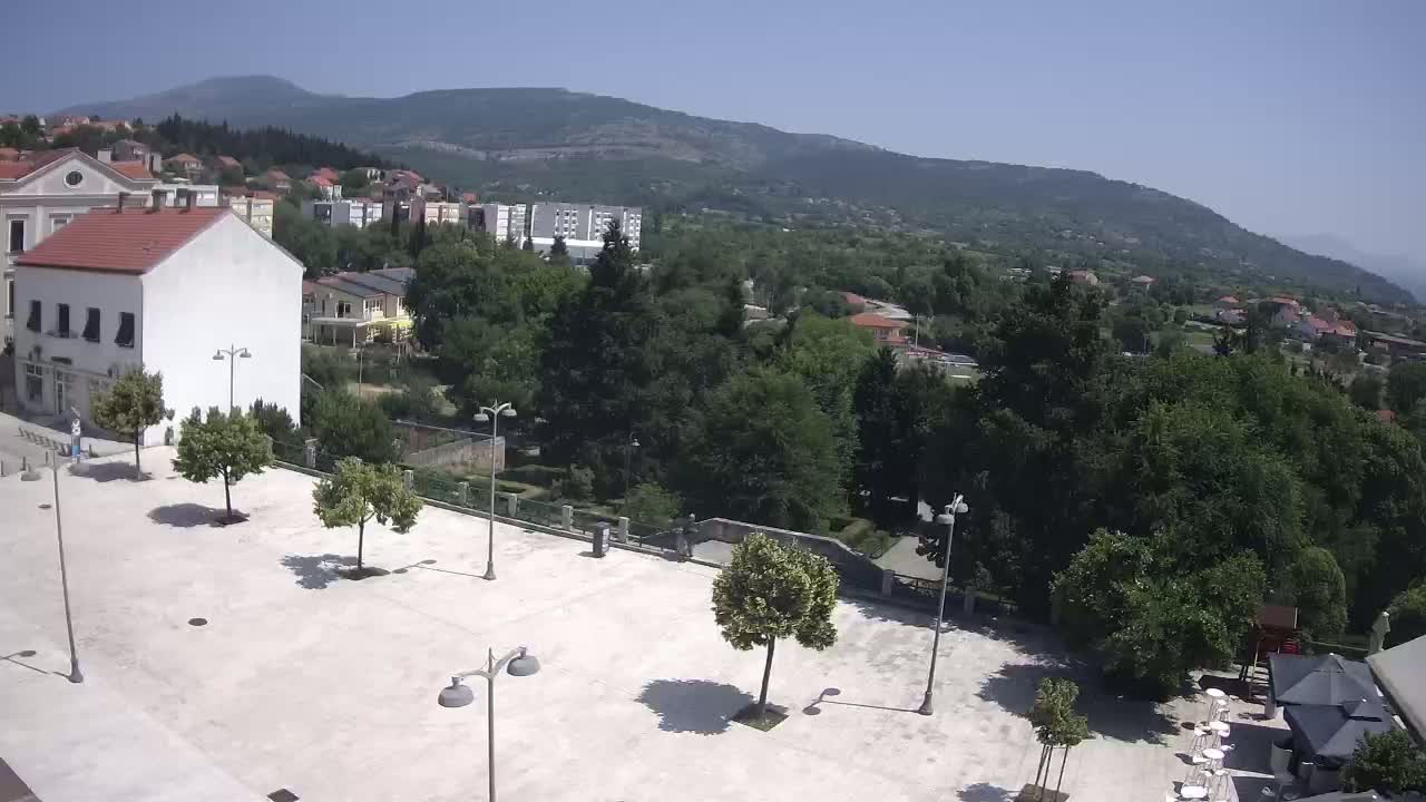 An empty town square with several trees and lampposts is situated in front of buildings, with a mountain range visible in the background under a clear, sunny sky.