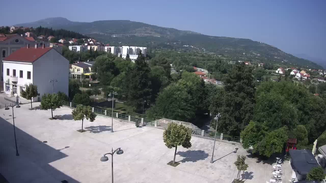 A mostly empty town square with several trees and lampposts, overlooks a green valley and distant mountains under a clear, sunny sky.