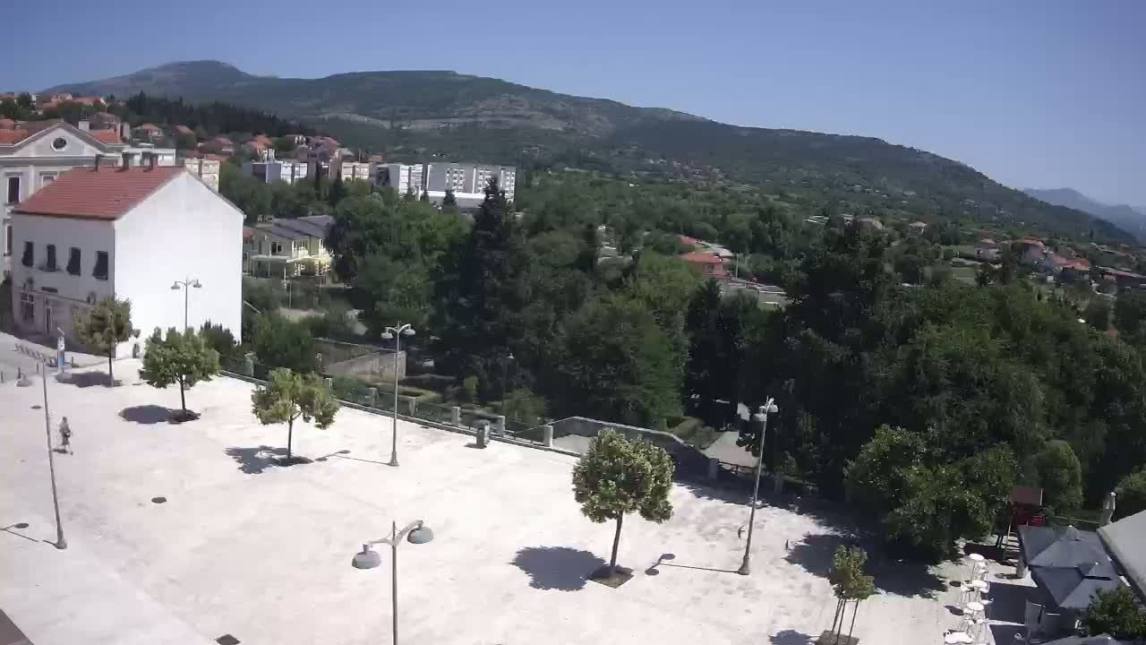 A mostly empty paved square is shown, with a few trees and light posts, overlooking a town nestled at the base of a mountain on a sunny day.