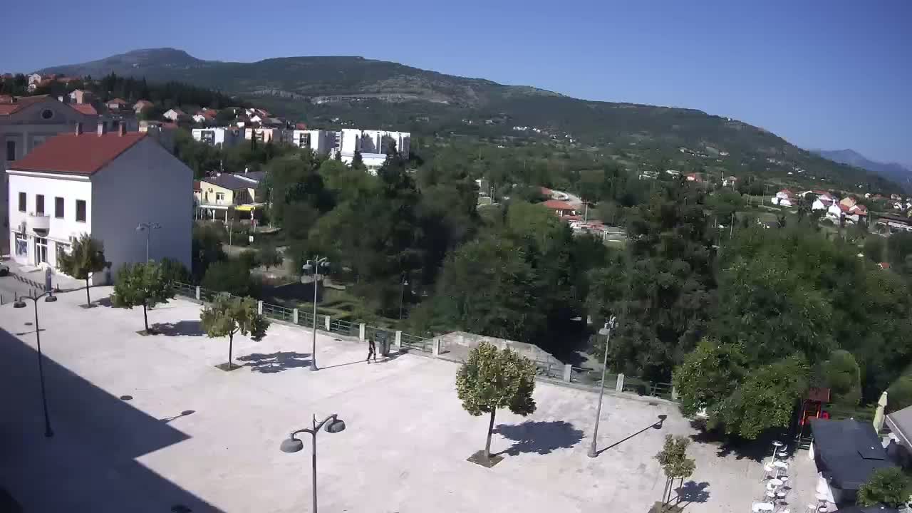 A paved town square with sparsely placed trees, overlooked by buildings and surrounded by lush greenery under a clear, sunny sky, with a mountain visible in the distance.
