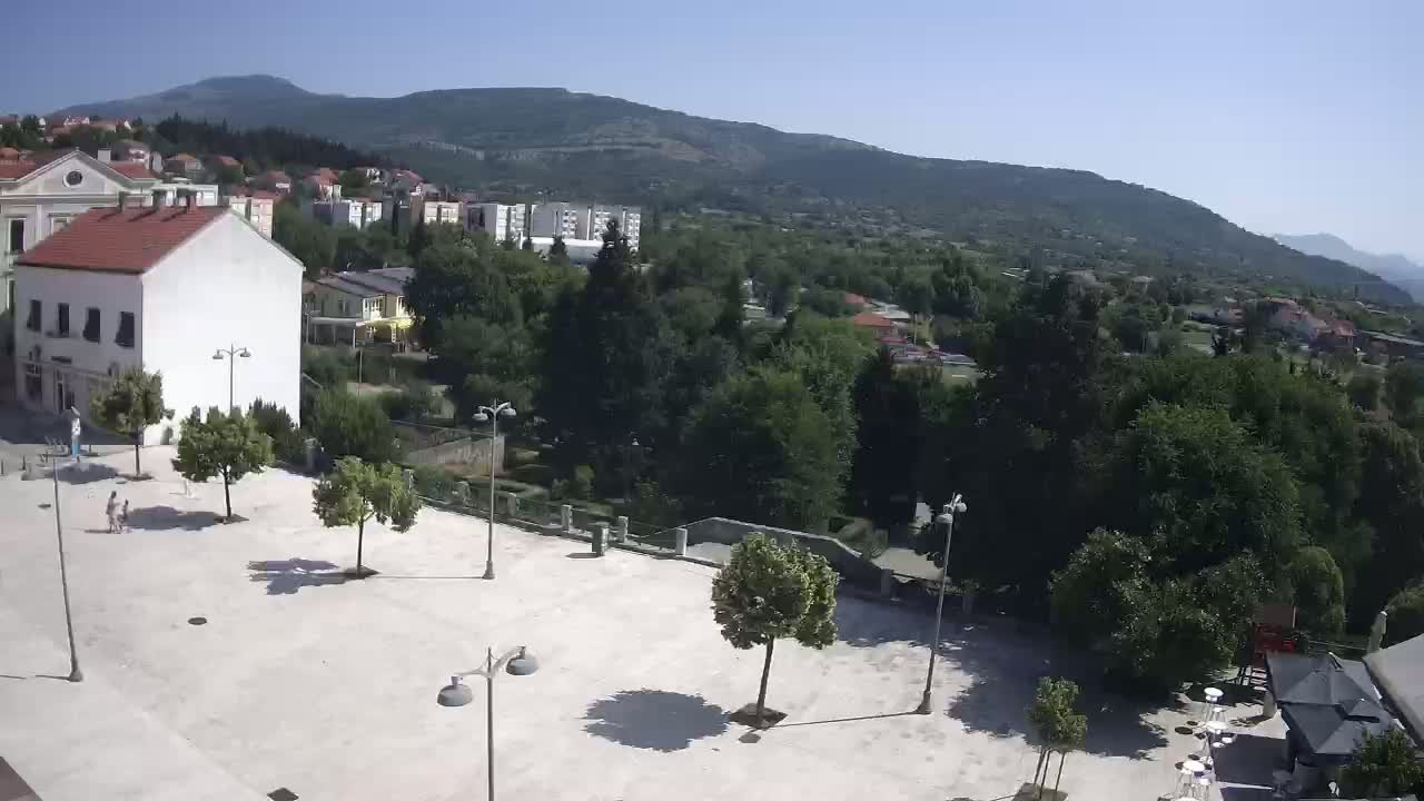 A sunny day overlooking a town square with several trees and buildings, backed by lush green hills and mountains.