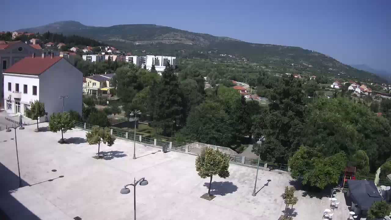 A mostly empty paved square is situated in front of buildings, overlooking a verdant valley and mountains under a clear, sunny sky.