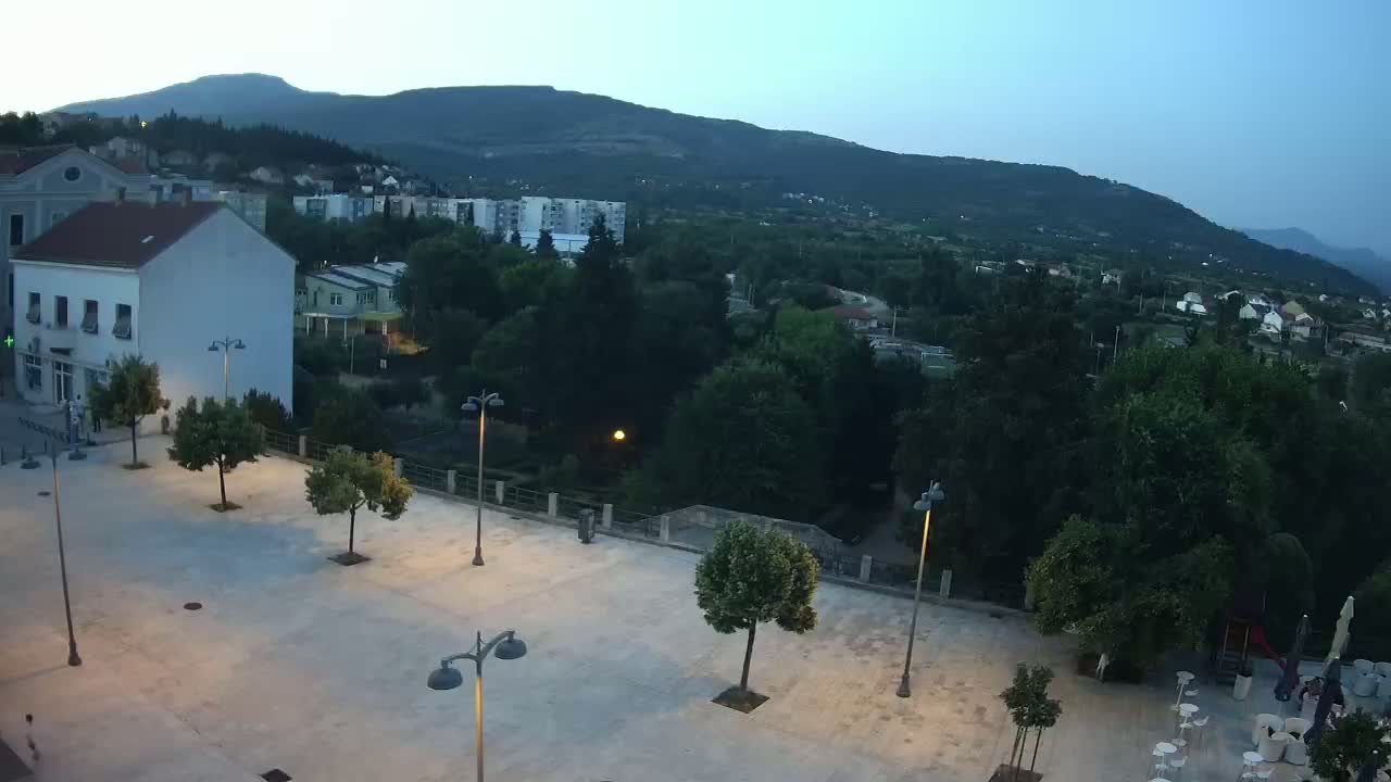 A mostly empty paved square is situated in front of buildings, overlooking a verdant valley and mountains under a clear, sunny sky.