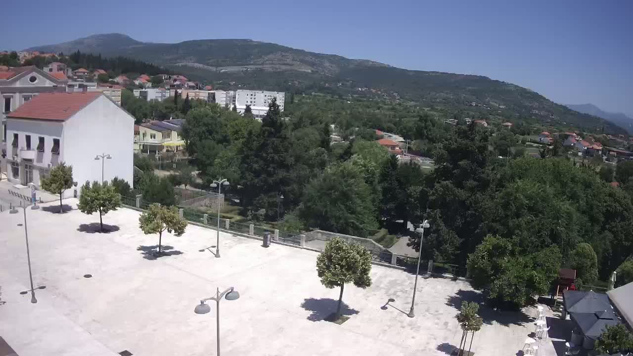 A mostly empty, light-colored paved square sits in front of buildings, surrounded by lush green trees and shrubs under a clear, sunny sky, with mountains in the background.