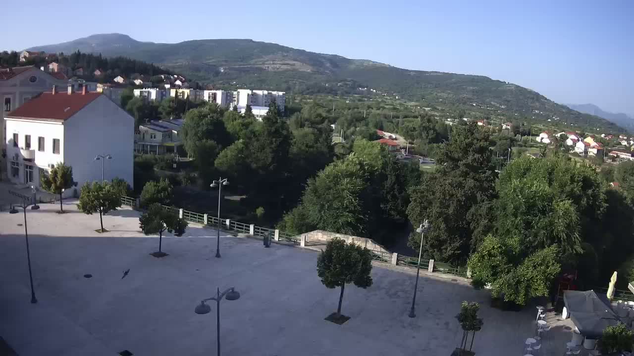 A mostly empty town square with several trees and lampposts, overlooking a river and a hillside town under a clear sky.