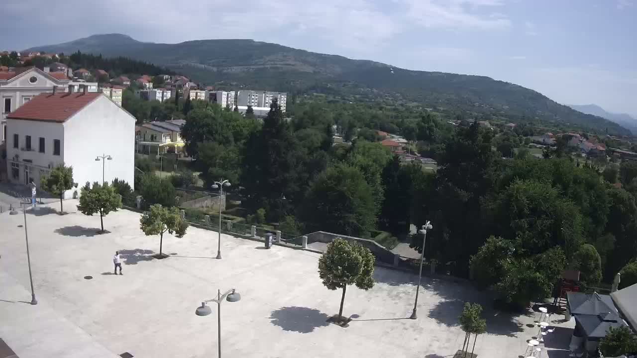 A paved town square with a few people and trees, overlooked by buildings and a mountain range under a mostly sunny sky.