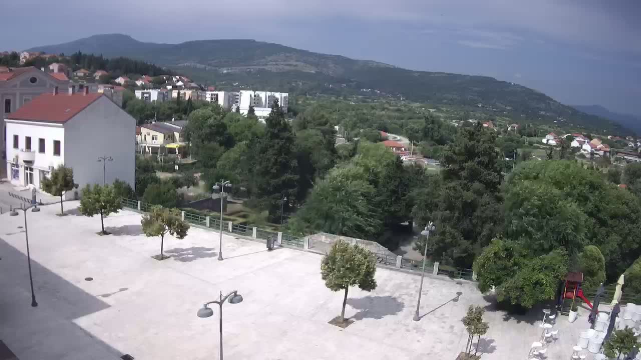 A mostly sunny, high-angle view of a town square with several trees and buildings, overlooking a lush green valley and distant mountains.