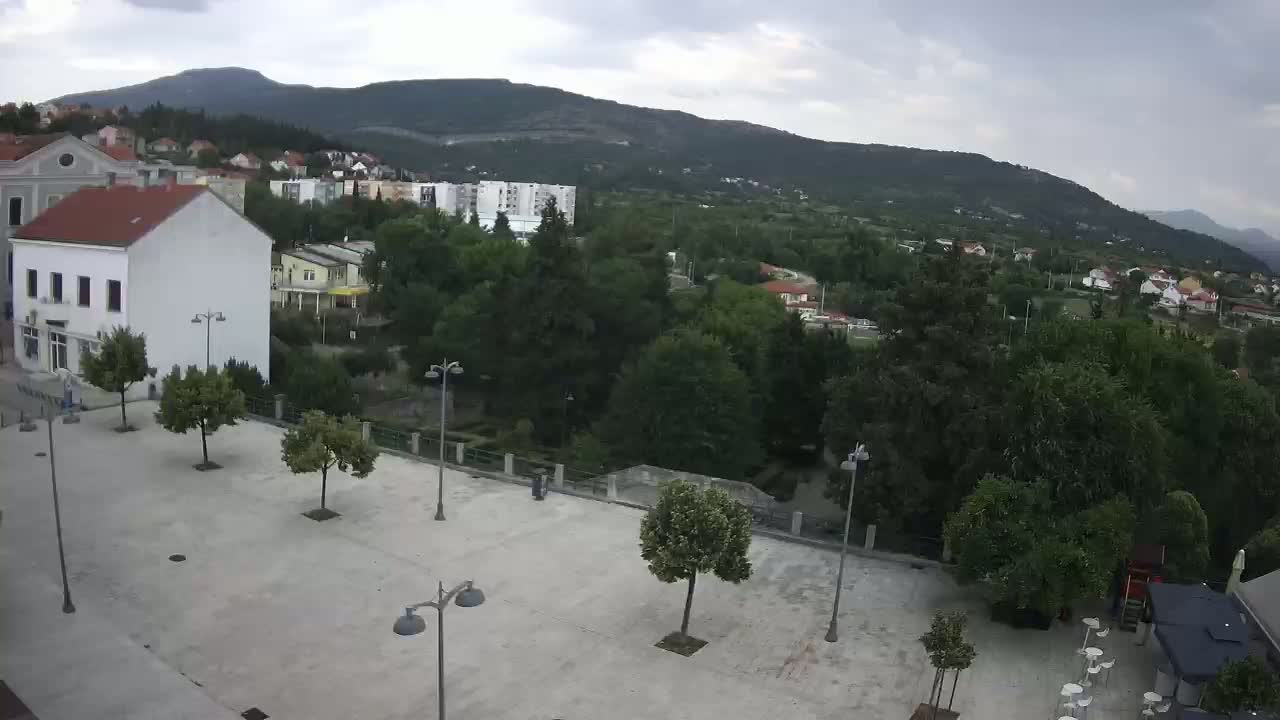 An overcast day reveals a town square with several trees and buildings, overlooked by a mountain range in the background.