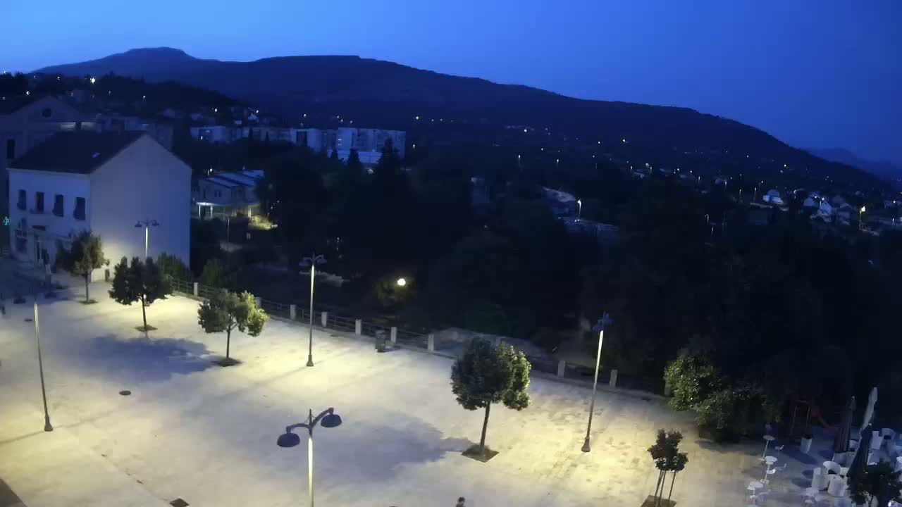 An empty, illuminated plaza at night overlooks a hilltop town under a twilight sky.