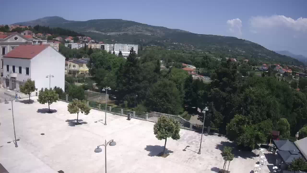 A mostly sunny view of a town square, surrounded by buildings and lush green trees, with mountains visible in the background.