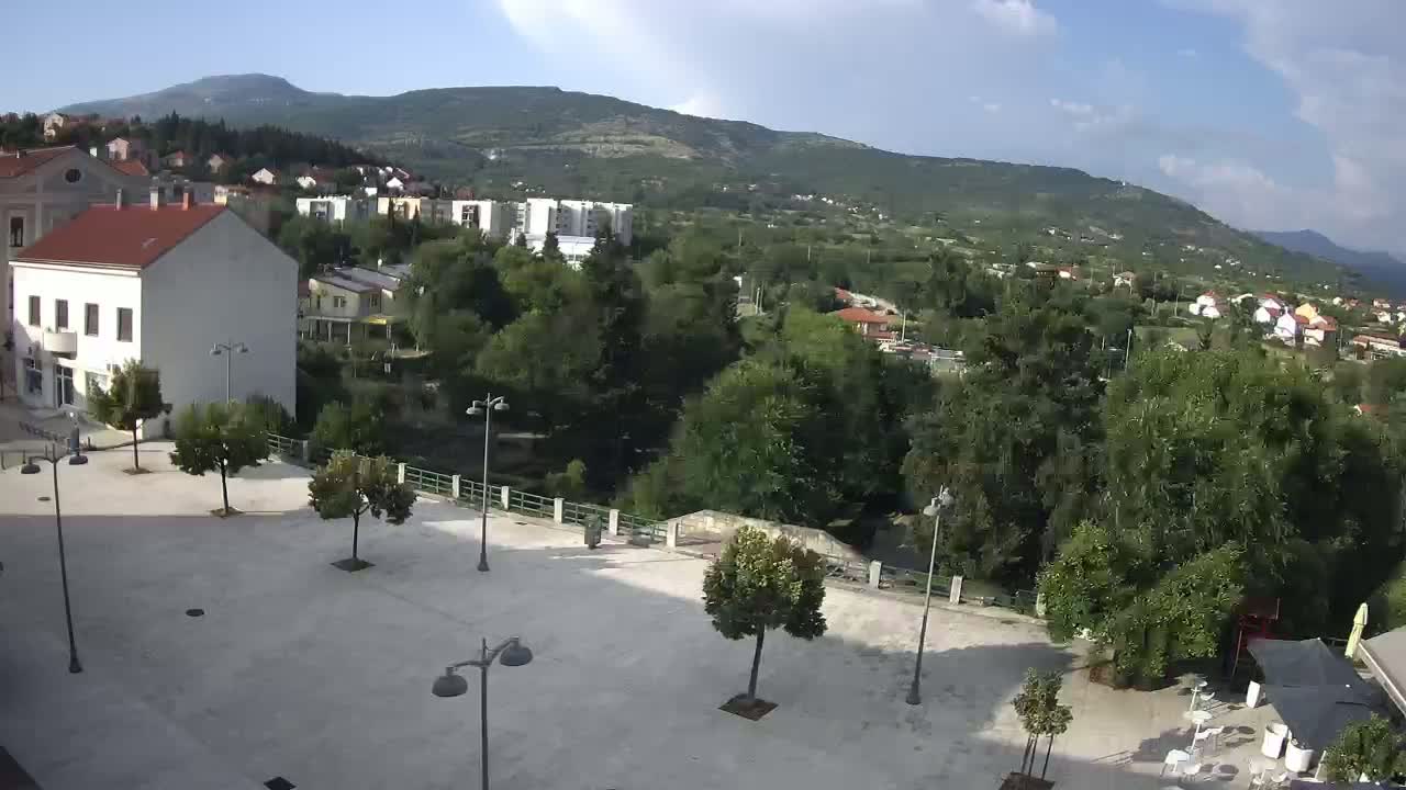 A mostly sunny day shows a town square with several trees and lampposts, overlooking a river, green hills, and residential buildings nestled against mountains in the distance.