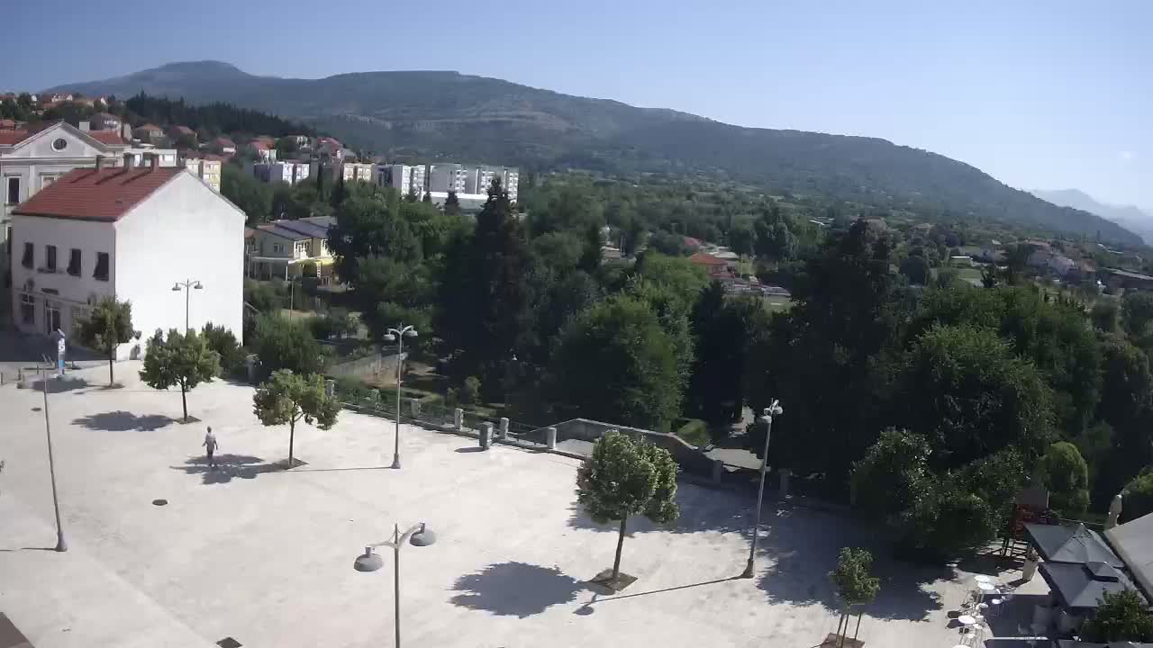 A mostly empty, light-colored square with several trees and buildings is visible, with a mountain range in the background under sunny skies.
