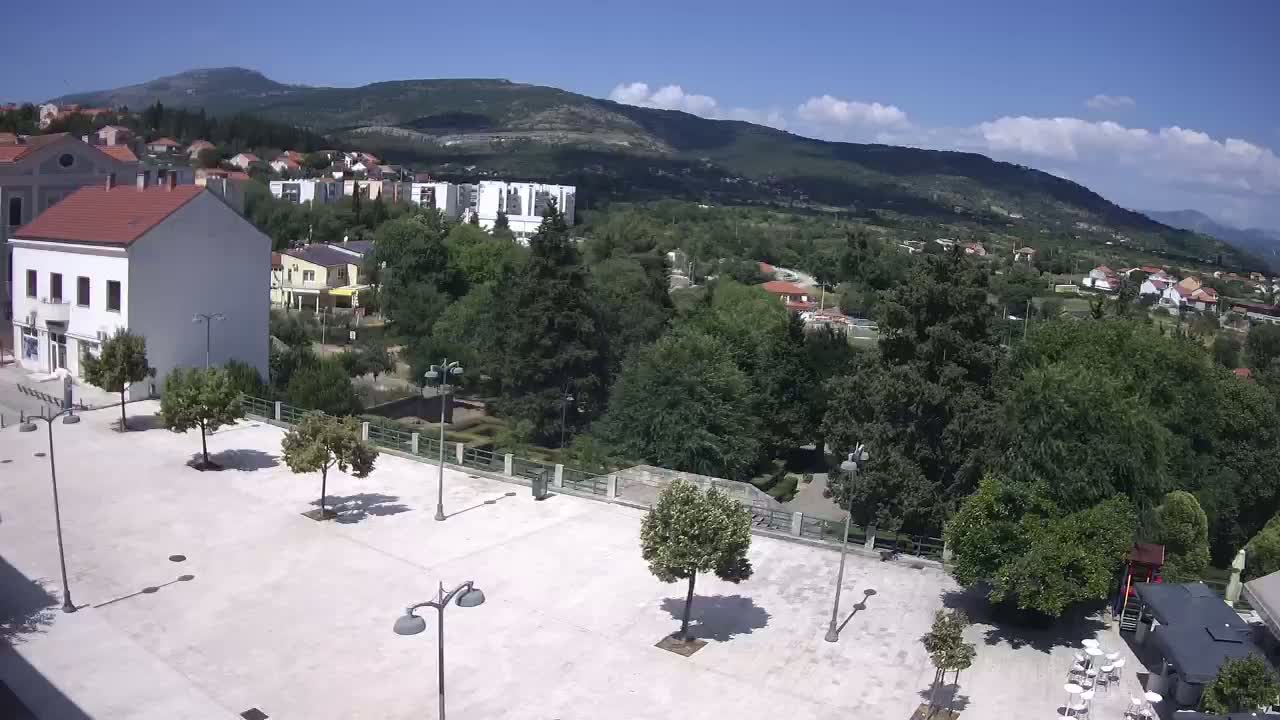 A mostly sunny day overlooks a town square with sparsely placed trees and buildings, and a mountain range in the distance.