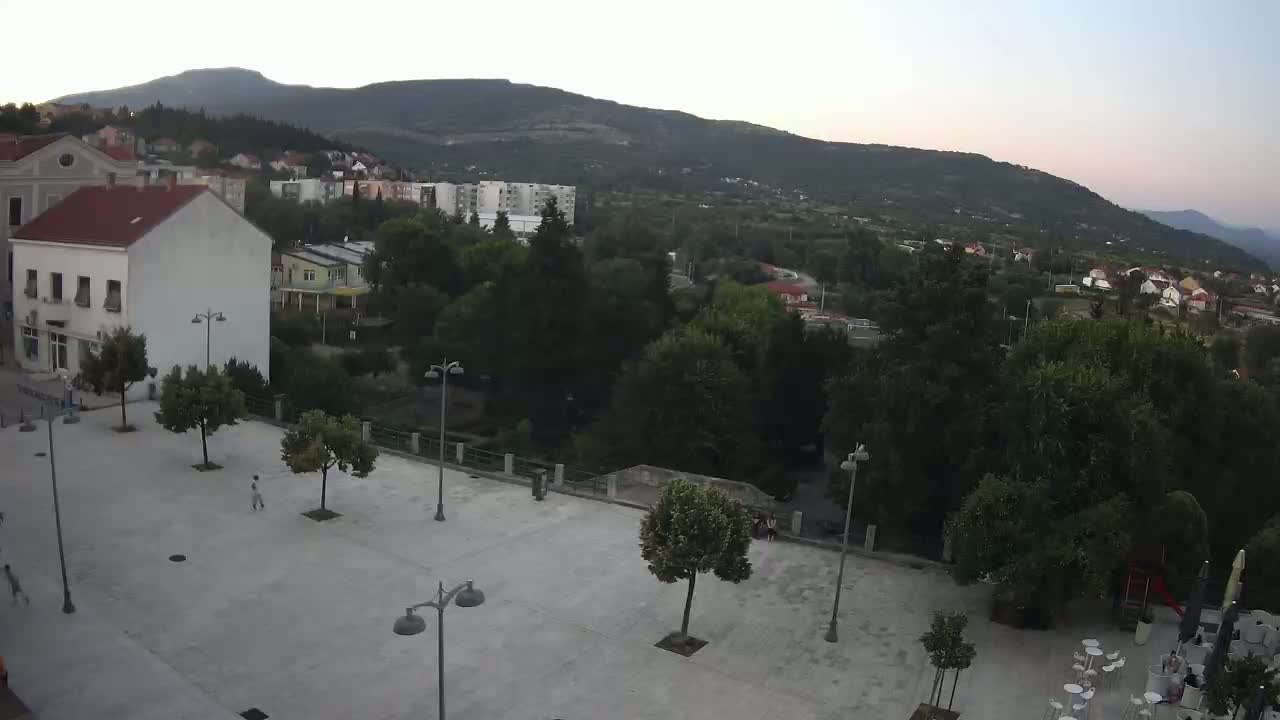 An empty town square with several trees and lampposts is situated in front of buildings and green hills under a clear evening sky.