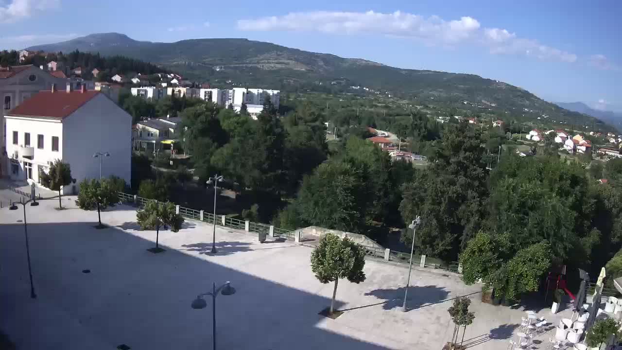 A paved plaza with several trees and lampposts, overlooks a town nestled among green hills under a mostly sunny sky.