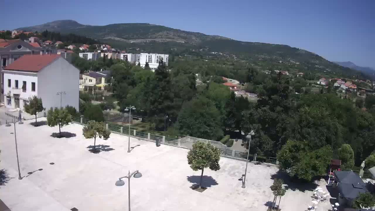 A paved town square with sparsely planted trees is situated below a hill in a sunny, clear-day setting.
