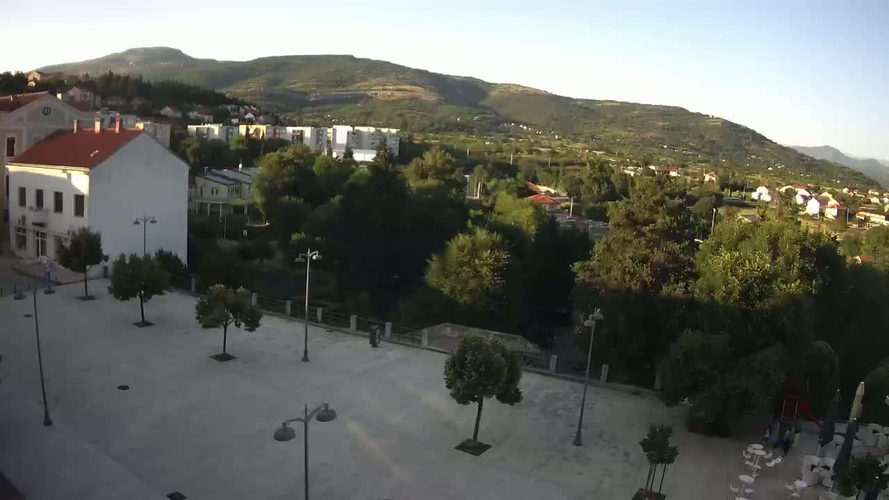A paved town square with several trees and lampposts is situated in front of buildings, overlooking a verdant valley and distant mountains under a clear sky.