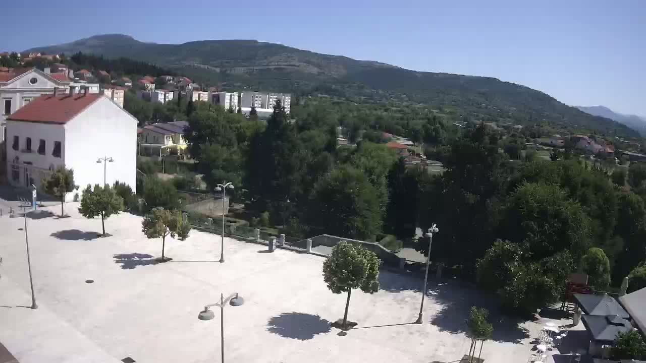 A mostly empty, light-colored paved square is shown, with several small trees and lamp posts, overlooking a town nestled in a valley below mountains on a sunny day.