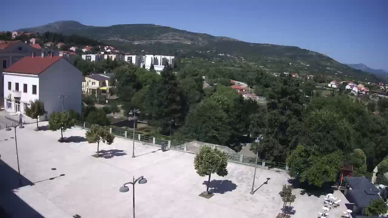 An empty paved square with several trees and lampposts overlooks a town nestled in a valley beneath a mountain on a clear, sunny day.