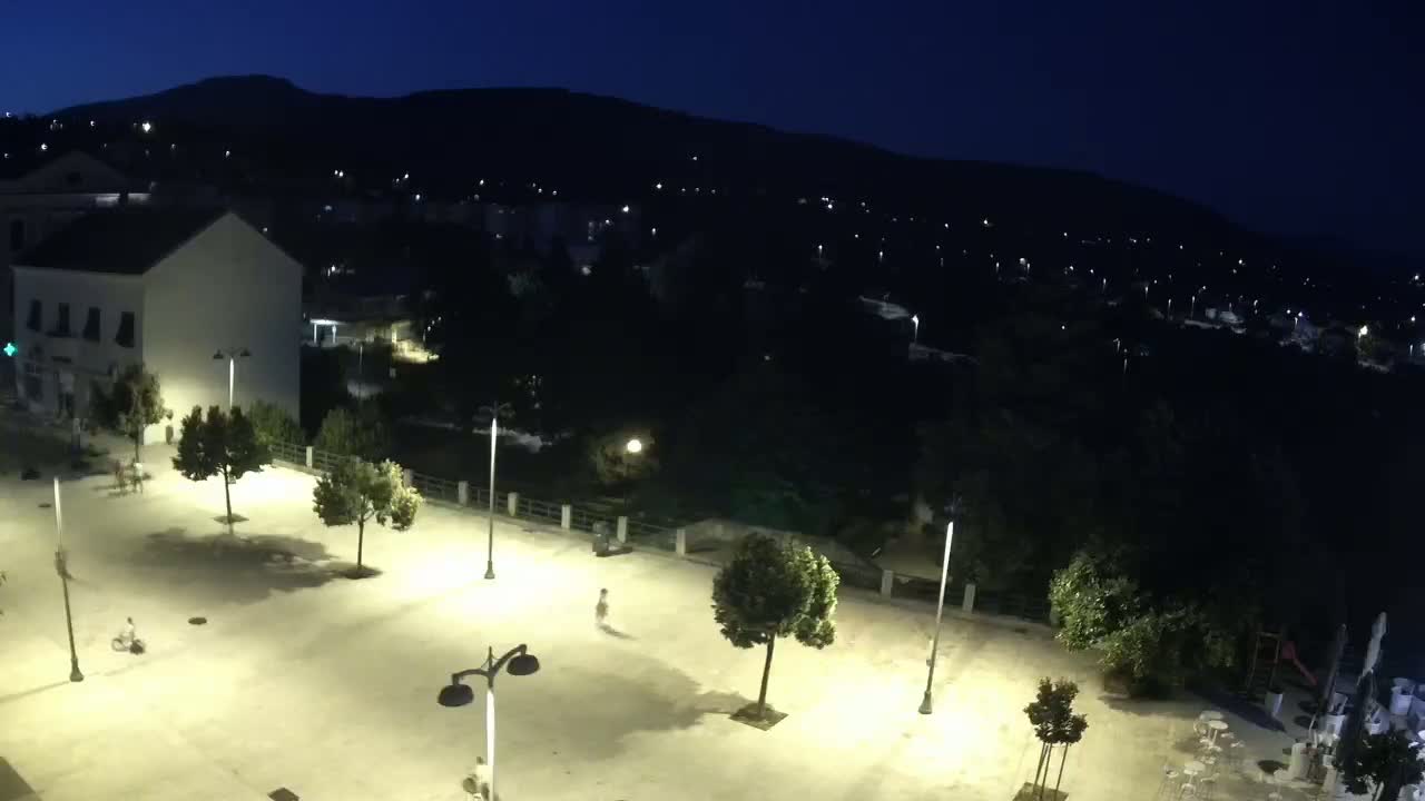 An empty, brightly lit town square at night is surrounded by trees and buildings, with a dark hill and sparsely lit town visible in the background under a clear, dark sky.