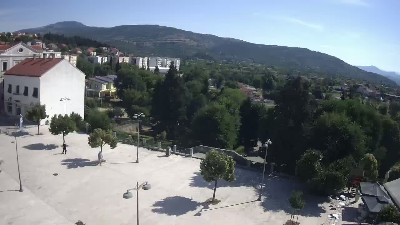 A paved town square with a few people and small trees, overlooked by buildings and a mountain range under a clear, sunny sky.
