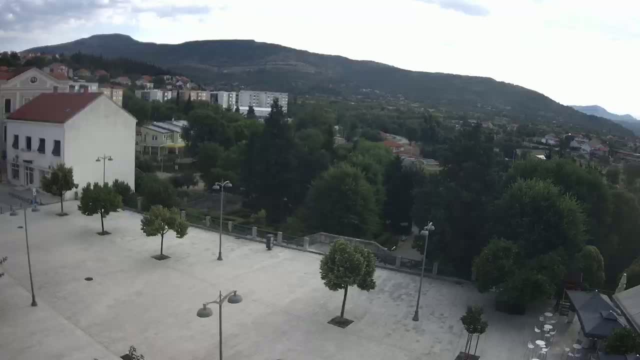 A paved town square with scattered trees and lampposts overlooks a verdant hillside town nestled beneath a mountain range on a partly cloudy day.