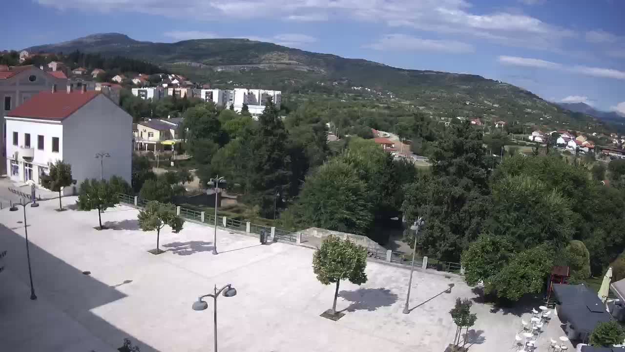 A paved town square with scattered trees and lampposts overlooks a verdant hillside town nestled beneath a mountain range on a partly cloudy day.