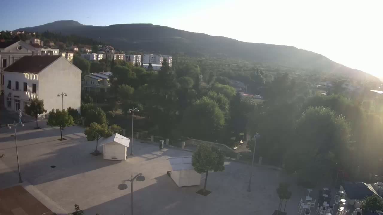 A paved town square with scattered trees and lampposts overlooks a verdant hillside town nestled beneath a mountain range on a partly cloudy day.